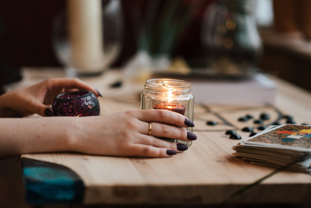 about-03 Hands with dark nails holding a lit candle, surrounded by tarot cards, creating a mystical and warm ambiance.