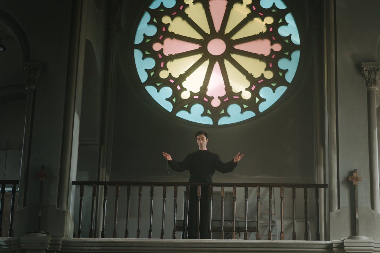 menu-02 A priest stands with open arms beneath a vibrant stained glass window inside a cathedral.